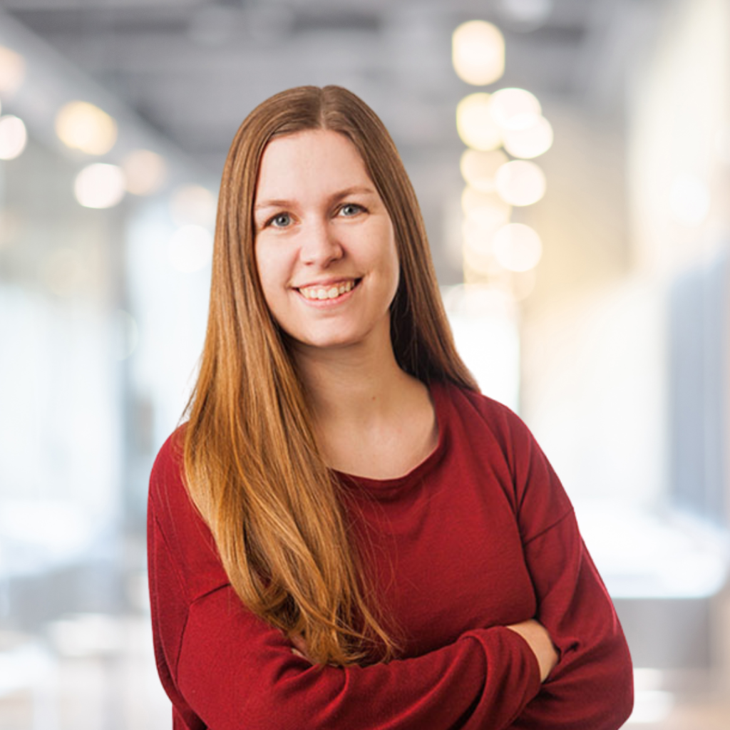 Junge Frau mit langen, glatten Haaren und einem freundlichen Lächeln, die in einem modernen Büro mit unscharfem Hintergrund steht. Sie trägt ein rotes Oberteil und hat die Arme vor der Brust verschränkt, was Selbstbewusstsein ausstrahlt.