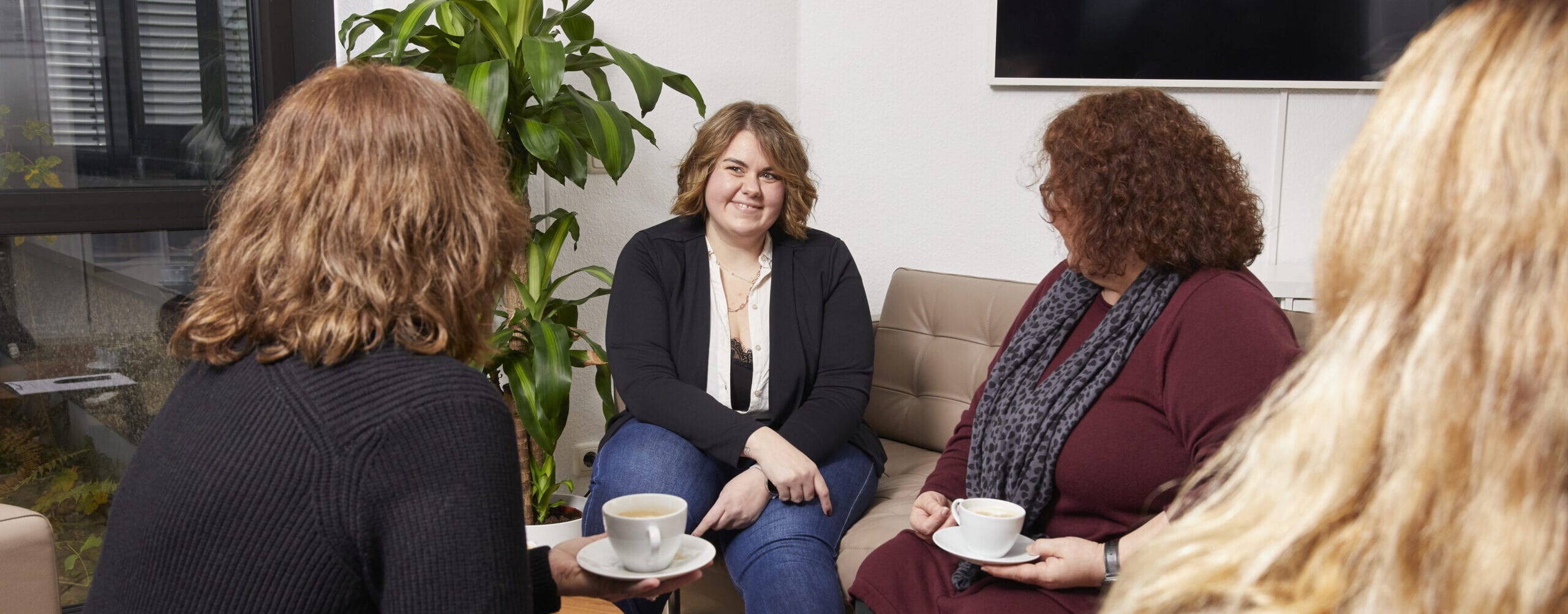 Vier Frauen sitzen in einem modernen Raum und unterhalten sich bei einer Tasse Kaffee. Eine Frau lächelt und blickt in die Runde, während die anderen aufmerksam zuhören. Im Hintergrund ist eine grüne Pflanze und ein Fernseher zu sehen, was eine einladende Atmosphäre schafft.
