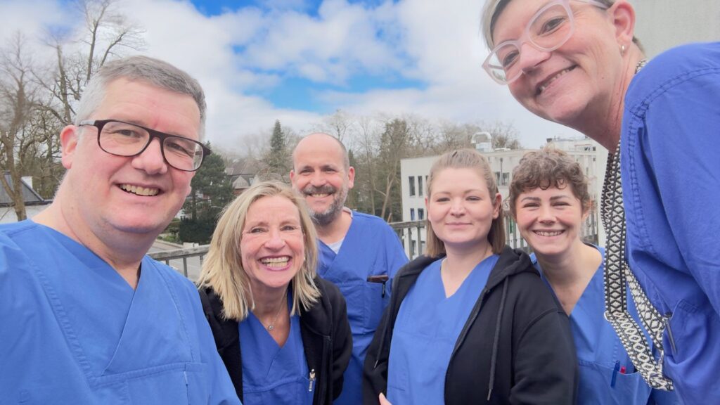 Gruppe von sechs medizinischen Fachkräften in blauen Kitteln lächelt in die Kamera, während sie auf einer Terrasse stehen, mit einem bewölkten Himmel im Hintergrund.