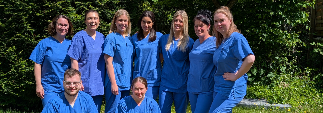 Gruppe von Fachkräften in blauen Arbeitskleidung, die im Freien stehen und lächeln. Die Aufnahme zeigt insgesamt neun Personen, die in einer grünen Umgebung posieren, was auf ein positives Arbeitsklima hinweist. Ideal für Themen rund um Gesundheitsberufe und Teamarbeit.