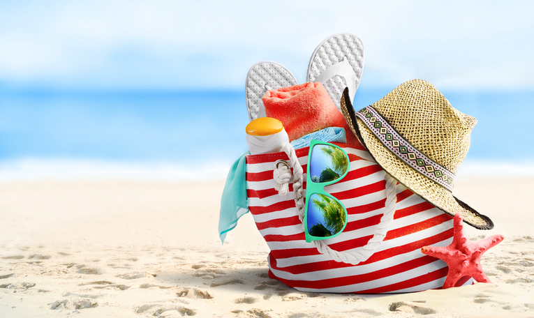Strandtasche mit Sonnenbrille, Sonnencreme, Handtuch und Flip-Flops auf einem Sandstrand, ideal für einen entspannten Urlaub am Meer.