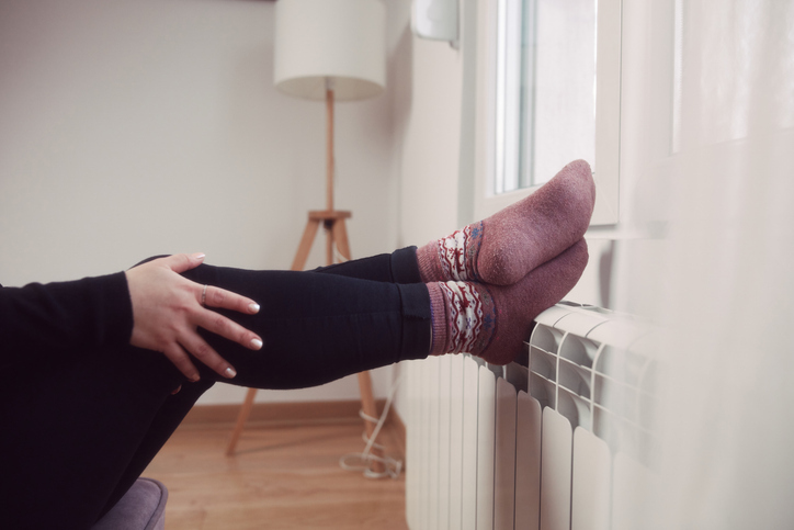 Eine Person mit bunten Socken sitzt entspannt mit den Füßen auf einem Heizkörper in einem hellen Raum, während sie eine gemütliche Atmosphäre genießt. Im Hintergrund ist ein Lampenständer sichtbar.