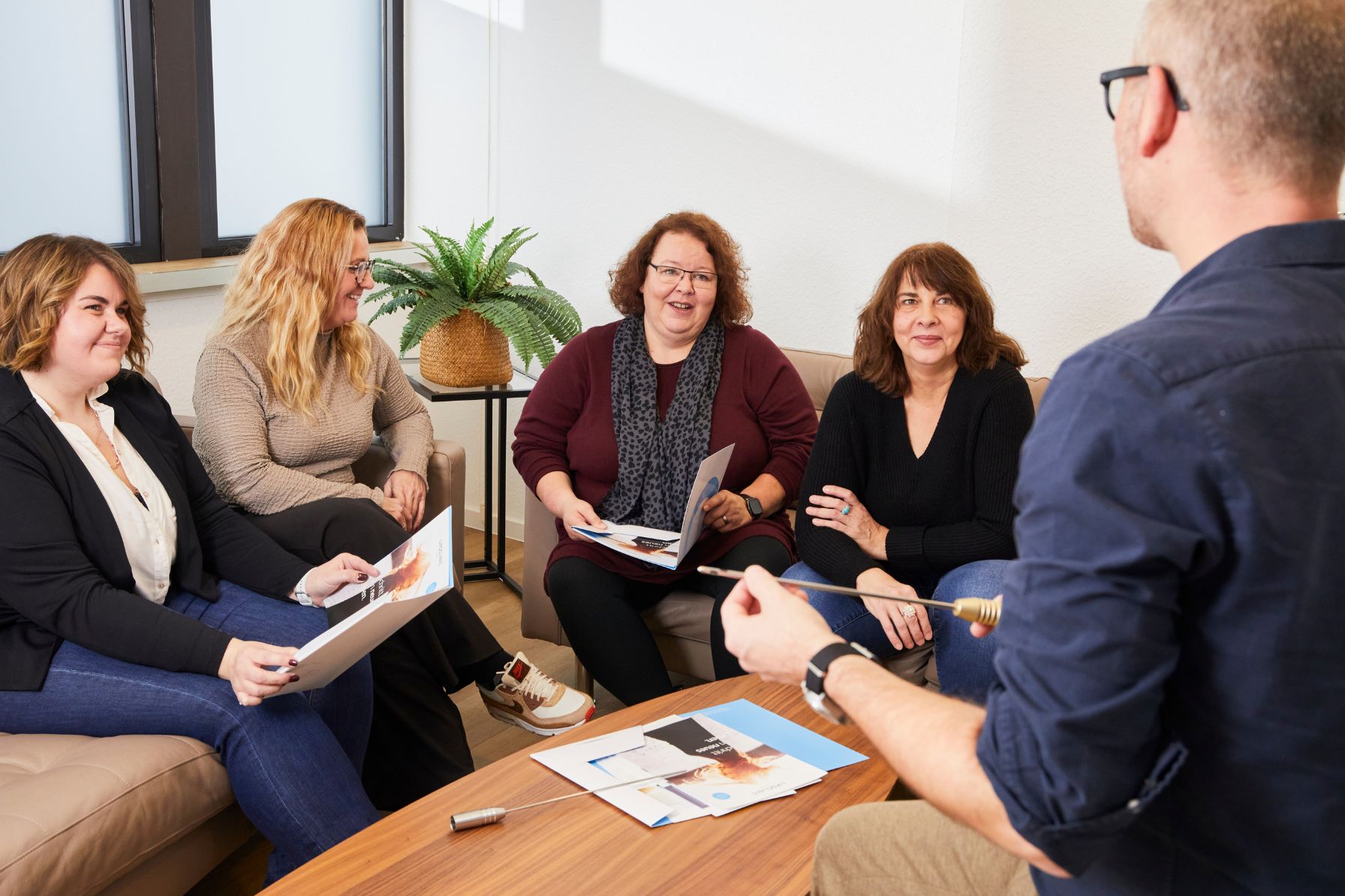 Fünf Personen in einem modernen Besprechungsraum, die an einem Tisch sitzen und sich aktiv austauschen. Sie halten Broschüren in der Hand und zeigen Interesse an der Präsentation. Eine Pflanze im Hintergrund sorgt für eine angenehme Atmosphäre.