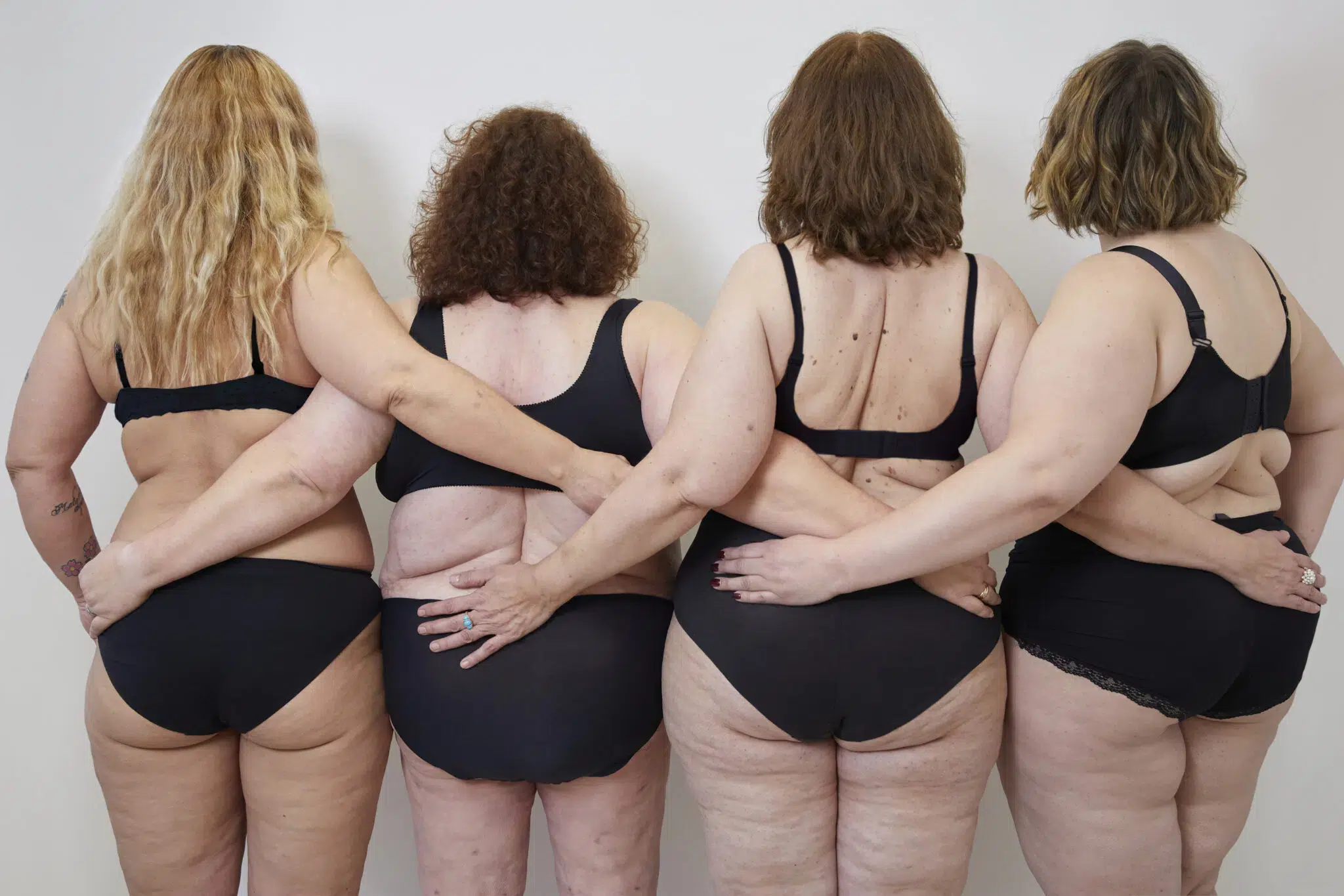 Vier Frauen mit unterschiedlichen Körperformen stehen Rücken an Rücken in schwarzer Unterwäsche und leiden an Lipoedem. Sie umarmen sich gegenseitig und zeigen Selbstvertrauen und Körperpositivität. Die Aufnahme hebt Vielfalt und Akzeptanz von verschiedenen Schönheitsidealen hervor.