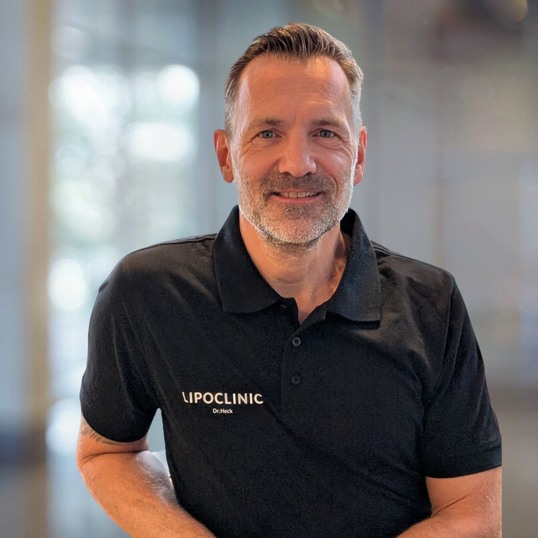 Porträt von Dr. Heck, einem Facharzt der Lipoclinic, der in einem schwarzen Poloshirt mit dem Logo der Klinik lächelt und eine freundliche Ausstrahlung hat. Der Hintergrund ist unscharf und vermittelt eine professionelle Atmosphäre.