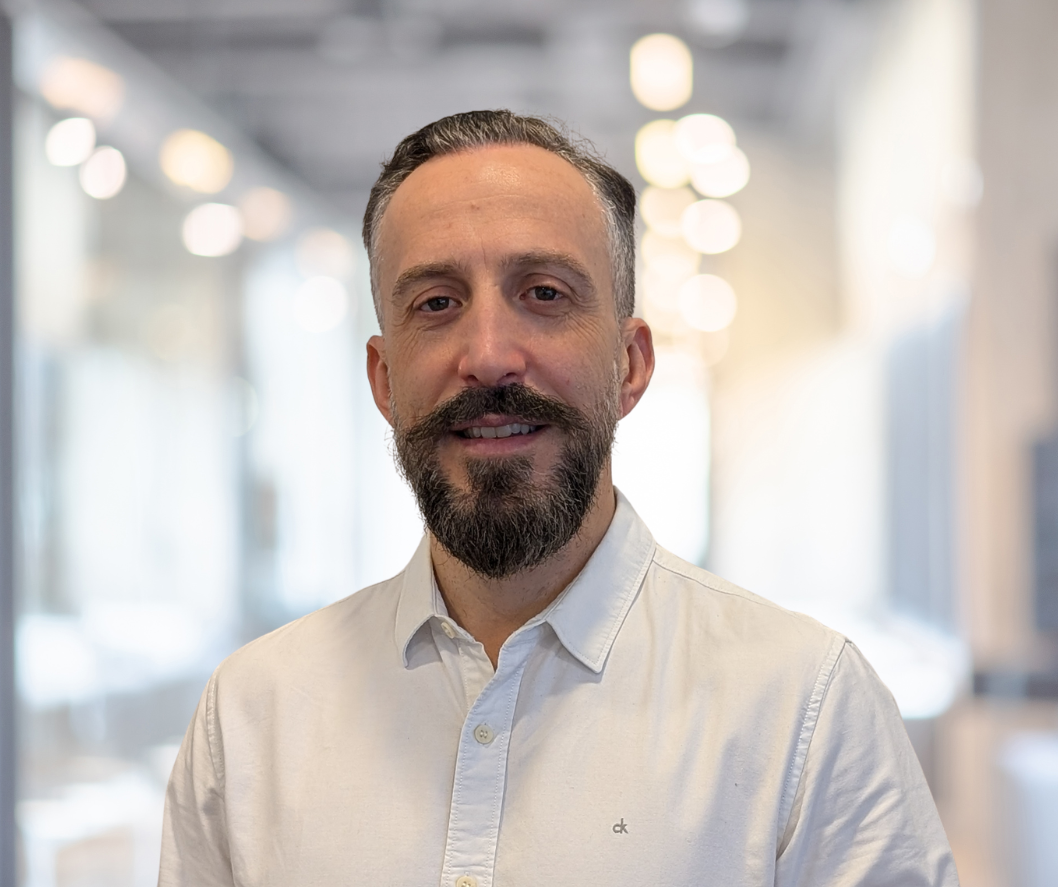 Portrait eines Mannes mit Bart und kurzen Haaren, der in einem modernen Büro steht. Er trägt ein helles Hemd und lächelt freundlich. Im Hintergrund sind unscharfe Lichter und Büromöbel sichtbar, was eine professionelle Atmosphäre schafft.