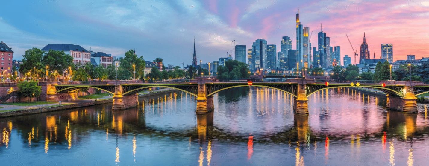 Panoramablick auf die Skyline von Frankfurt am Main bei Sonnenuntergang, mit der Alten Brücke im Vordergrund, die über den Fluss Main führt, umgeben von modernen Wolkenkratzern und historischen Gebäuden.