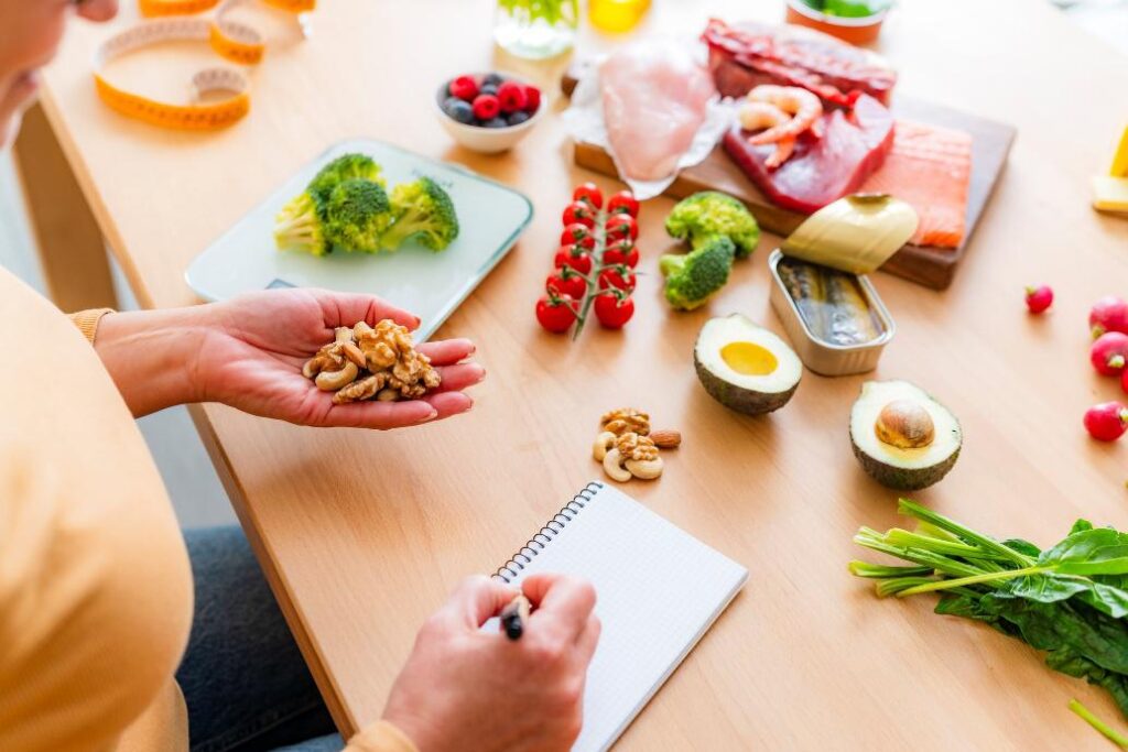 Eine Person hält eine Mischung aus Nüssen in der Hand und schreibt in ein Notizbuch, während auf dem Tisch frisches Gemüse, Obst und verschiedene proteinreiche Lebensmittel wie Fisch und Fleisch liegen. Die Szene vermittelt einen gesunden Lebensstil und die Planung einer ausgewogenen Ernährung.