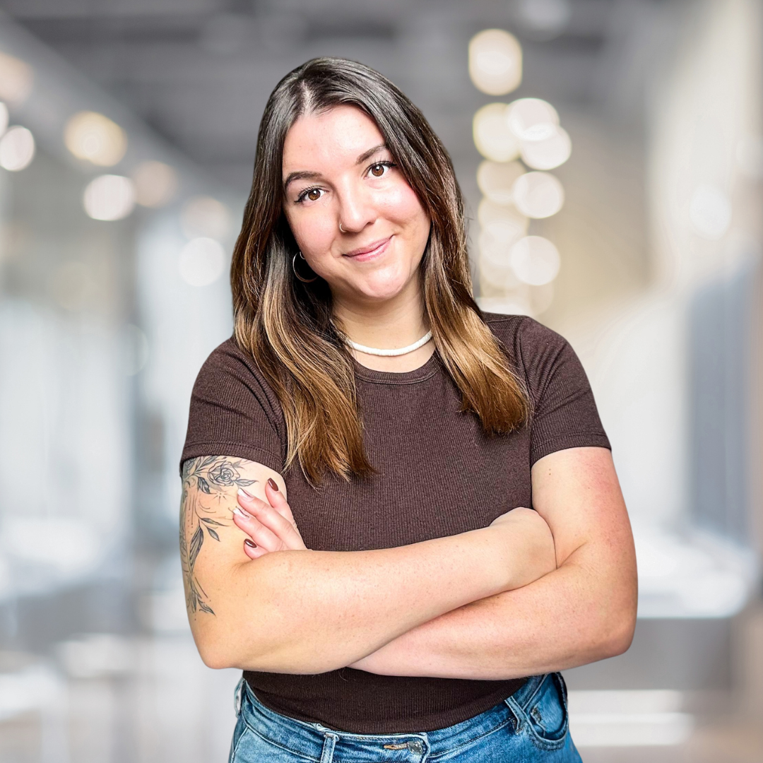 Porträt einer jungen Frau mit langen, lockigen Haaren und Tattoos, die in einem modernen Büro steht. Sie trägt ein braunes T-Shirt und hat die Arme vor der Brust verschränkt. Der Hintergrund ist unscharf und zeigt eine helle, einladende Büroumgebung mit weichen Lichtakzenten.