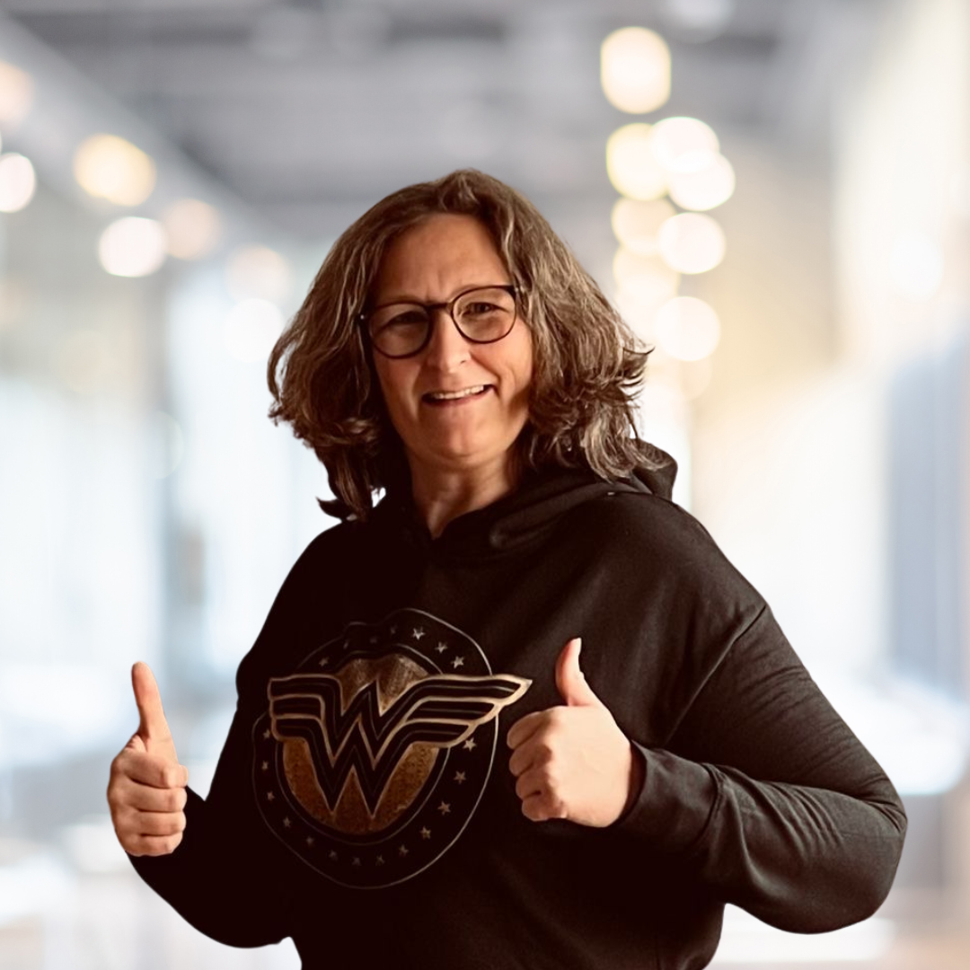 Frau mit lockigem Haar und Brille, die in einem schwarzen Hoodie mit Wonder Woman-Logo steht und Daumen hoch zeigt, vor einem modernen, hellen Hintergrund.
