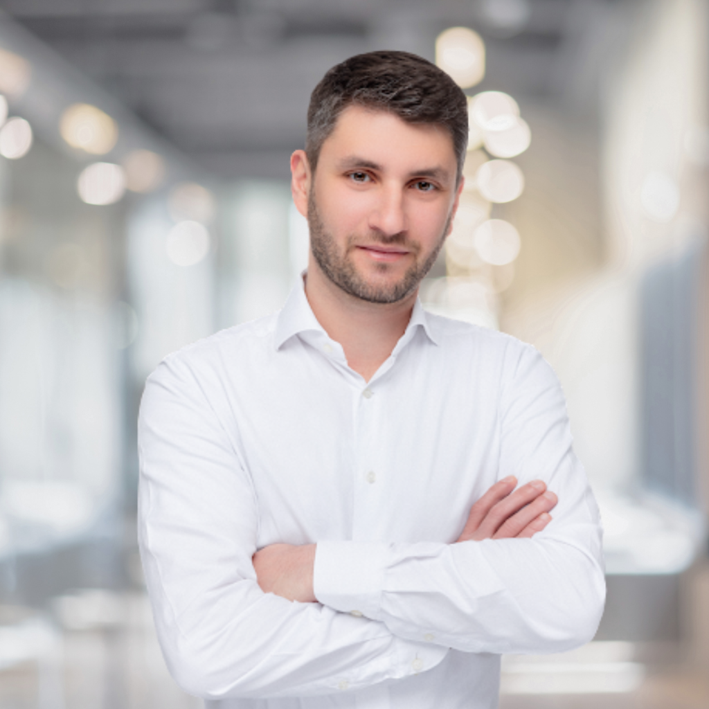 Professioneller Mann in weißem Hemd mit verschränkten Armen, stehend in einem modernen Büro mit unscharfem Hintergrund, der eine einladende und selbstbewusste Ausstrahlung vermittelt.