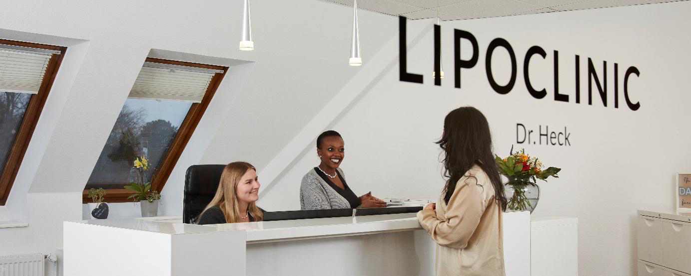 Empfangsbereich der Lipoclinic Dr. Heck mit freundlichen Mitarbeiterinnen, die eine Patientin beraten. Helle, moderne Einrichtung mit Pflanzen und dekorativen Elementen.