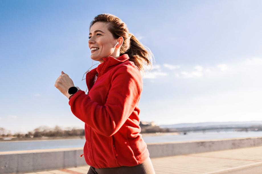 Frau in roter Jacke joggt am Wasser entlang, trägt Kopfhörer und Fitnessuhr, strahlt Freude und Energie aus, bei klarem Himmel und sonnigem Wetter.
