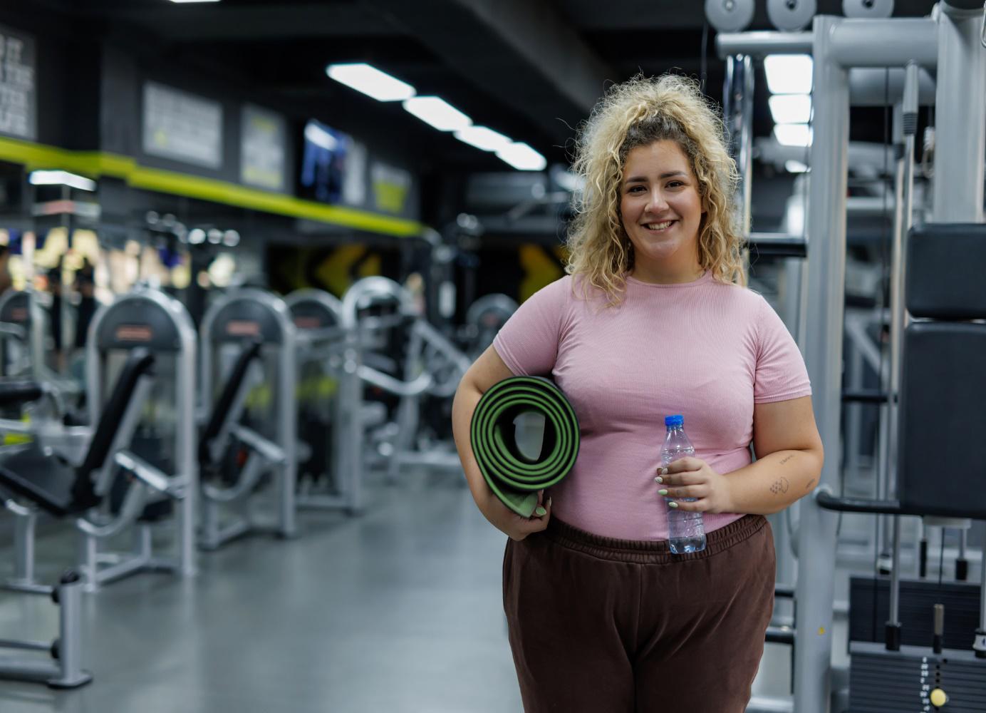 Junge Frau im Fitnessstudio mit Sportmatte und Wasserflasche, bereit für das Training. Hintergrund zeigt moderne Fitnessgeräte und eine motivierende Umgebung.
