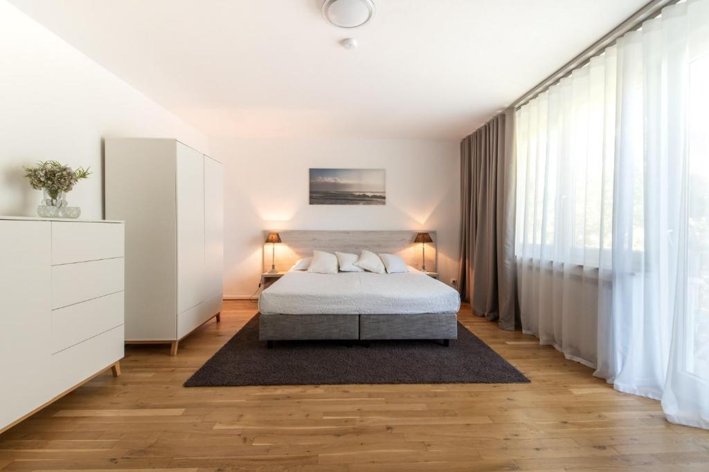 Helles und modernes Schlafzimmer mit einem Doppelbett, einem großen Fenster mit Vorhängen, einem stilvollen Nachttisch mit Lampe und einem minimalistischen Kleiderschrank. Der Fußboden aus Holz und der Teppich sorgen für eine gemütliche Atmosphäre.