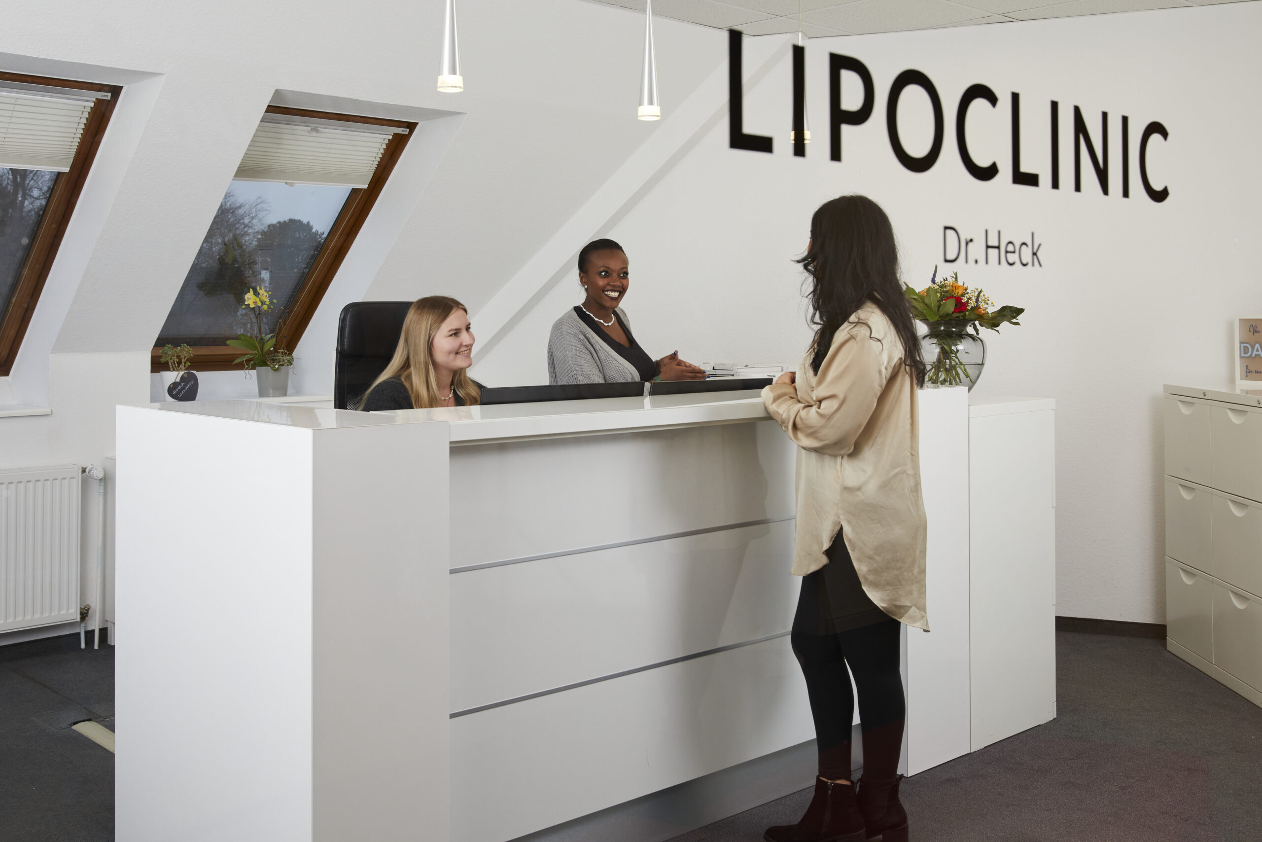 Empfangsbereich der Lipoclinic Dr. Heck mit freundlichem Personal und einer Patientin, die Informationen erfragt. Helle, moderne Einrichtung und Pflanzen sorgen für eine einladende Atmosphäre.