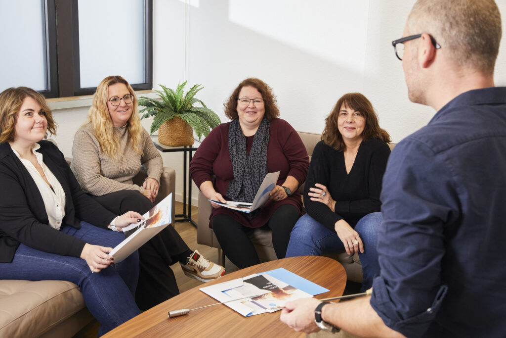 Teammeeting in einem modernen Büro: Fünf Personen, darunter vier Frauen und ein Mann, sitzen in einem gemütlichen Raum und diskutieren. Sie halten Unterlagen in den Händen und schauen aufmerksam zu dem Mann, der ihnen gegenübersteht. Eine Pflanze im Hintergrund sorgt für eine angenehme Atmosphäre.
