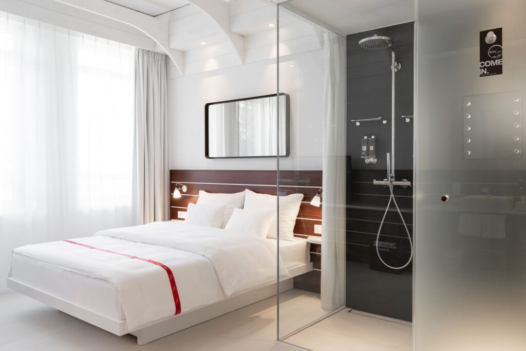 Helles und modernes Hotelzimmer mit einem Doppelbett, weißen Bettwäsche und einer eleganten, gläsernen Dusche. Die Wände sind in neutralen Farben gehalten, während die Fenster mit transparenten Vorhängen versehen sind, die für viel Tageslicht sorgen. Ideal für einen entspannten Aufenthalt.