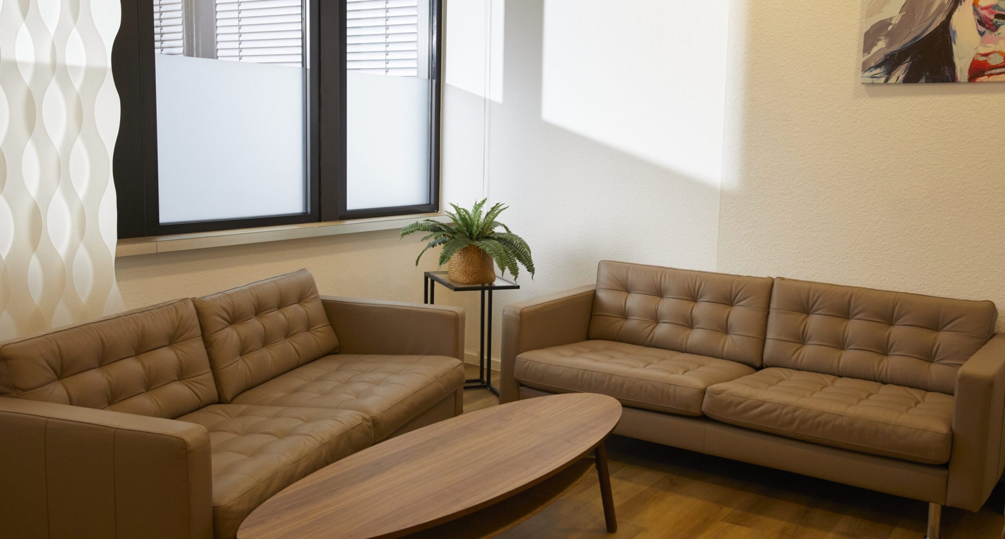 Moderne Lounge-Einrichtung mit zwei beigen Sofas, einem runden Holztisch und einer Zimmerpflanze in einem hellen Raum mit großen Fenstern und dekorativen Vorhängen. Beratung Lipödem.