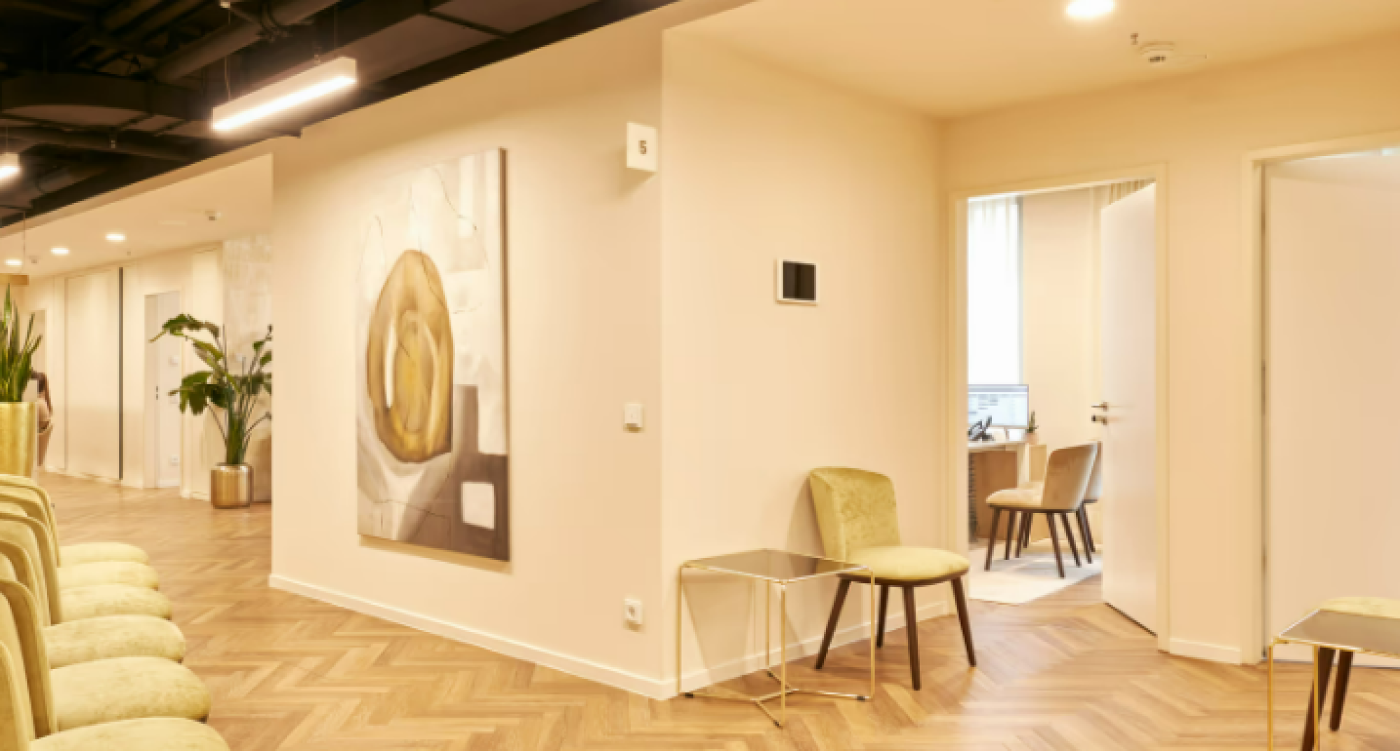 Moderne Büroumgebung mit stilvollen Sitzplätzen, einem Kunstwerk an der Wand und einem hellen, einladenden Raum. Der Boden ist mit Fischgrätmuster verlegt, und es gibt Pflanzen zur Dekoration.