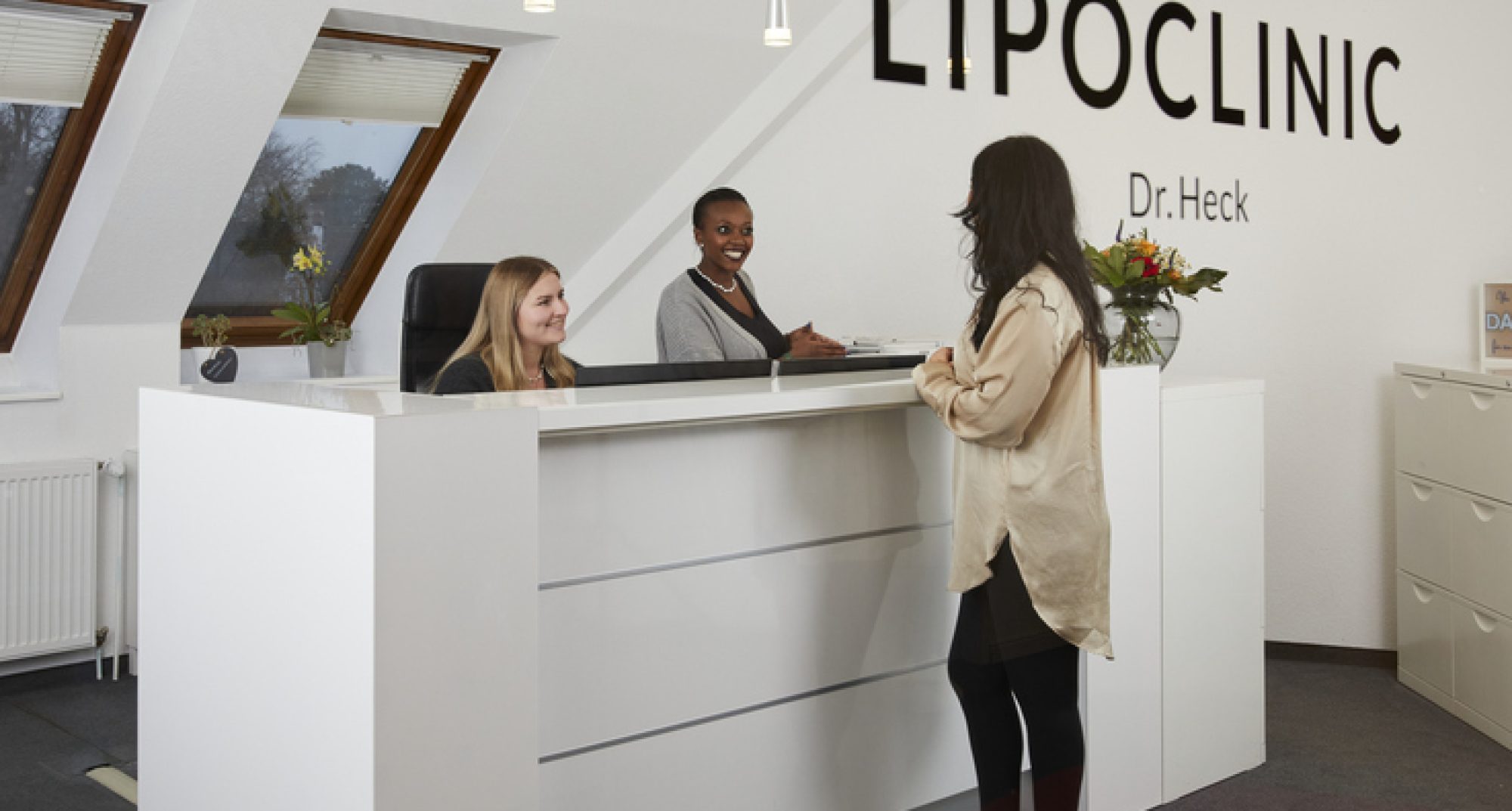 Empfangsbereich der Lipoclinic Dr. Heck mit zwei Mitarbeiterinnen, die eine Patientin begrüßen, passend zum Thema Kontakt.